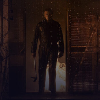 Man in a mask walking out of a burning building with an ax in his hand