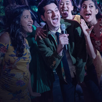 Four people performing, singing and dancing while one of them, in the middle, holds the microphone.