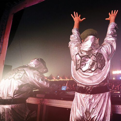 N.A.S.A from the back performing at a concert. On the left, one of the members at the mixing table. On the right the other member with his arms up in the air.