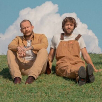 Artists Penny & Sparrow sitting on the grass with a blue cloudy sky in the background