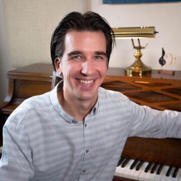 Headshot of the composer Matt Janszen sitting at a piano and smiling